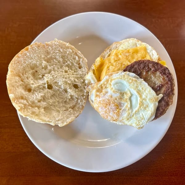 A breakfast sandwich with sausage, egg, and cheese on a bun, served open-faced on a white plate.