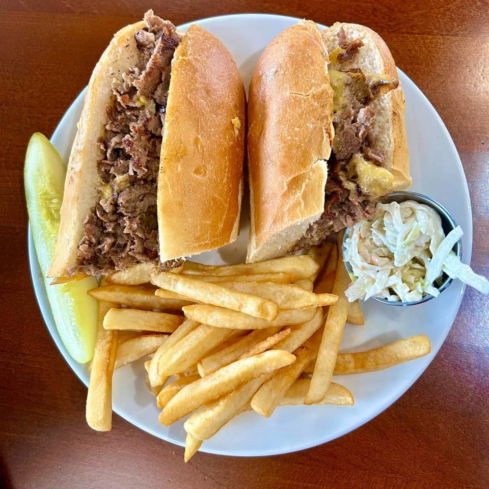 A cheesesteak sandwich, cut in half, with fries, a pickle spear, and coleslaw on a white plate.