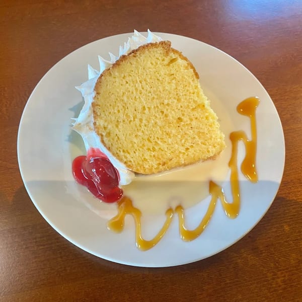 A slice of yellow cake with whipped cream, cherry topping, and caramel drizzle on a white plate.