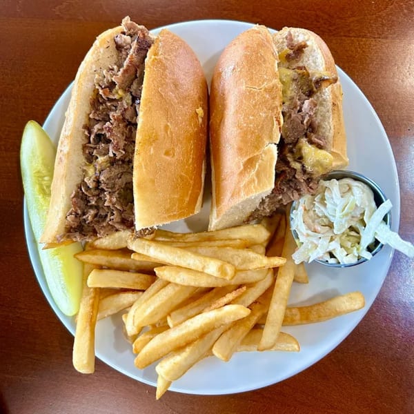 Cheesesteak sandwich cut in half, served with golden fries, a pickle spear, and coleslaw.
