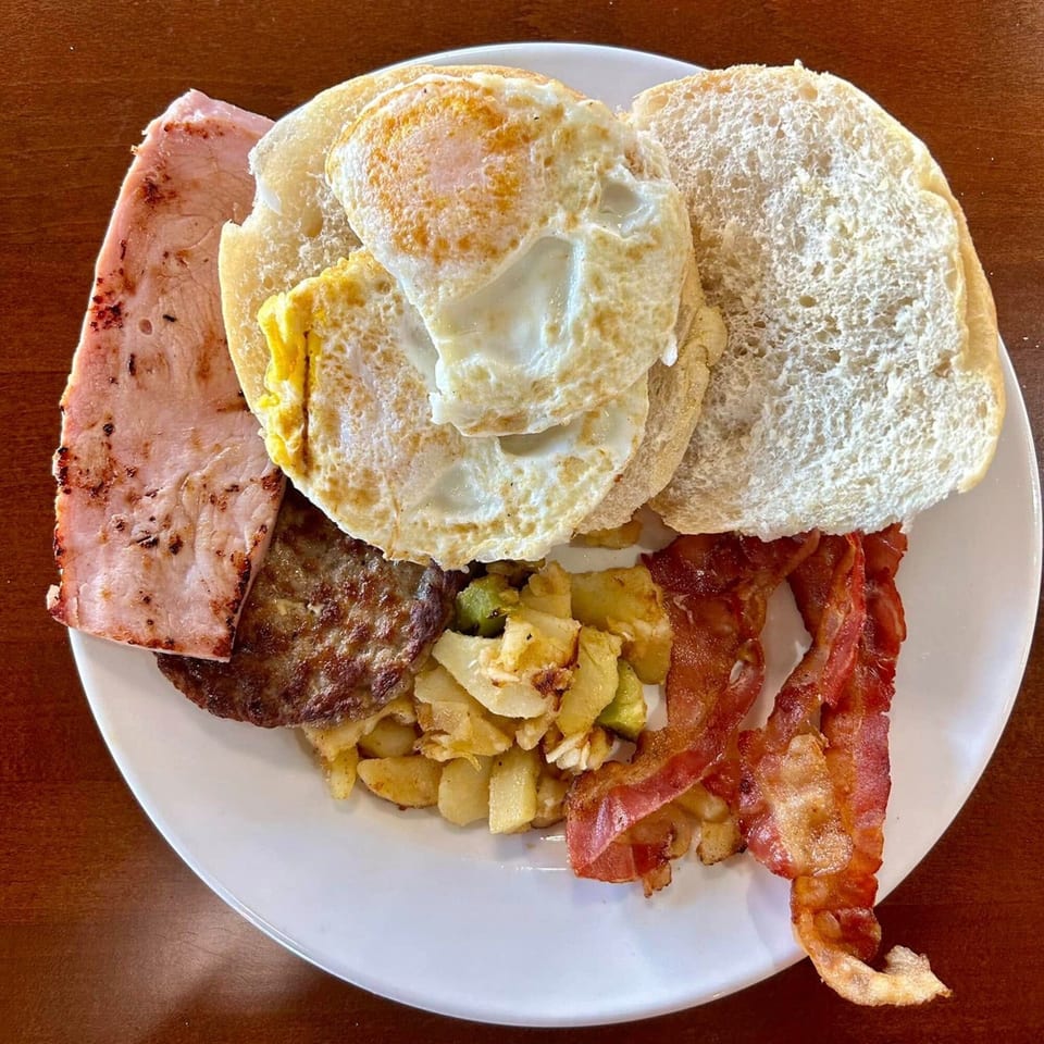 A breakfast platter with fried eggs, bacon, ham, sausage, home fries, and a bun.